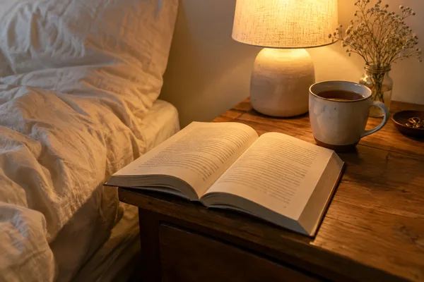An open book and a mug of tea on a wooden bedside table lit by a lamp.